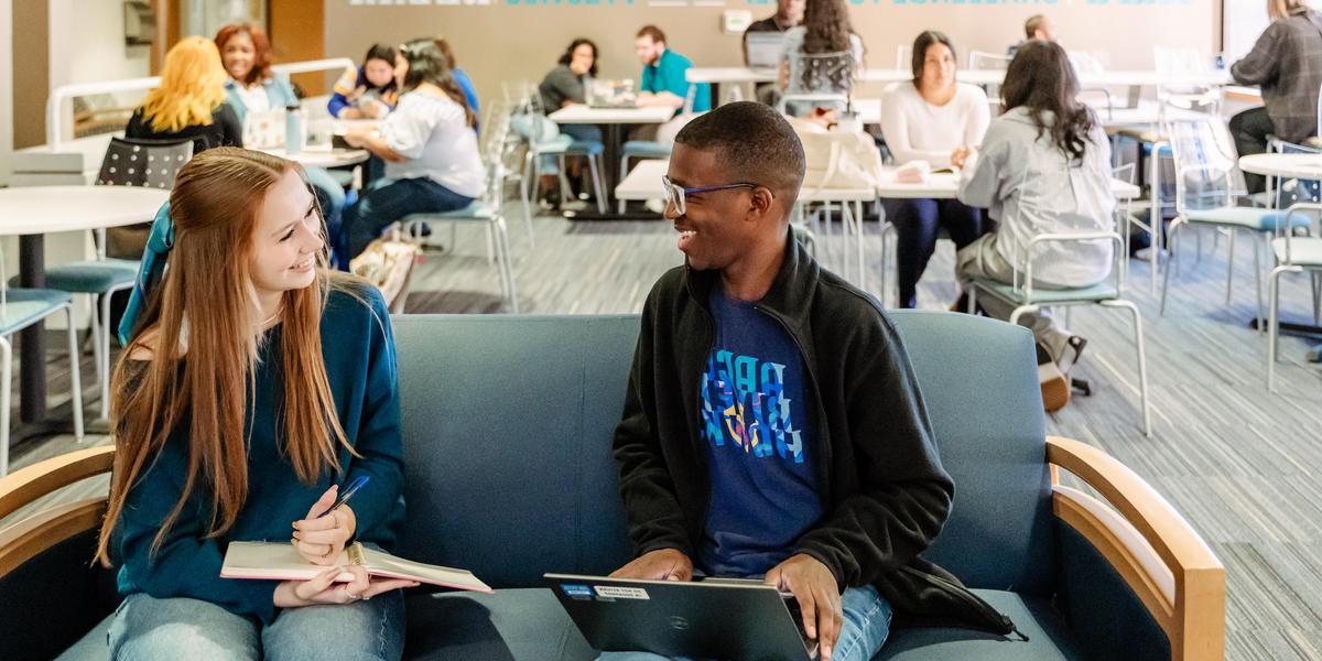 Two students, one on a laptop, one writing in a notebook, talk in the Testing Center Lounge